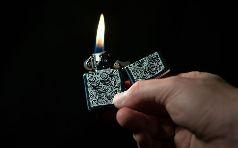 hand holding a metal lighter with ornate, swirling engravings and a small flame against a dark background.
